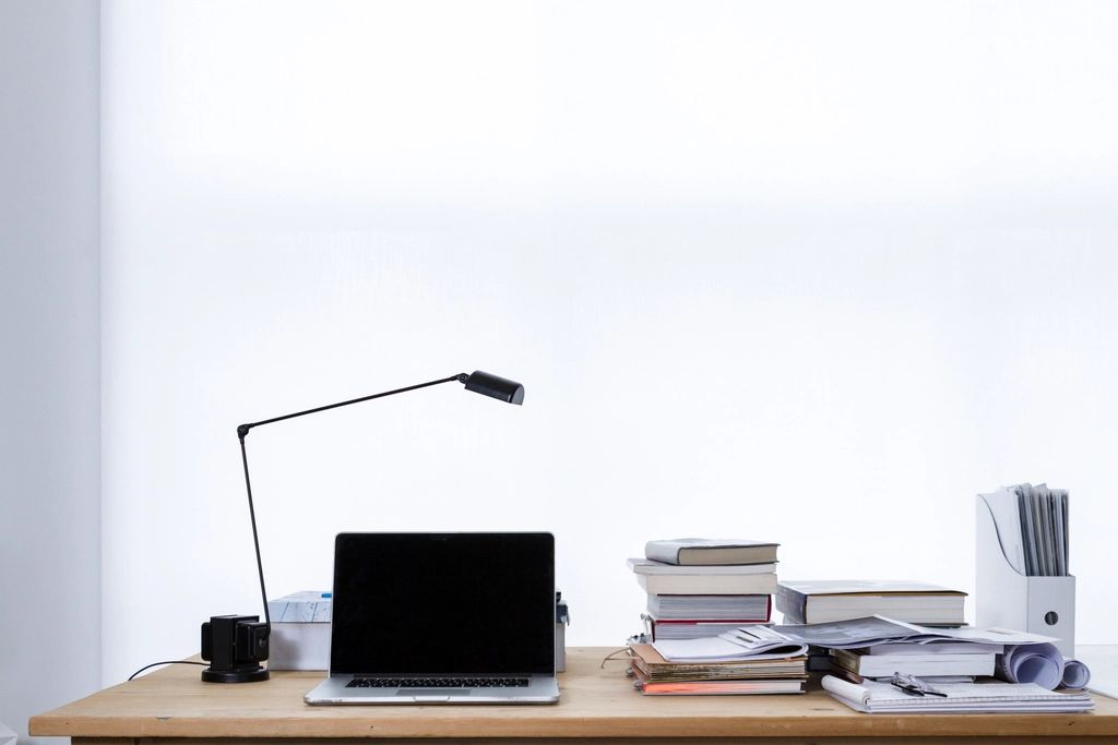 A tidy desk with a lamp, a closed laptop and stacks of books and papers