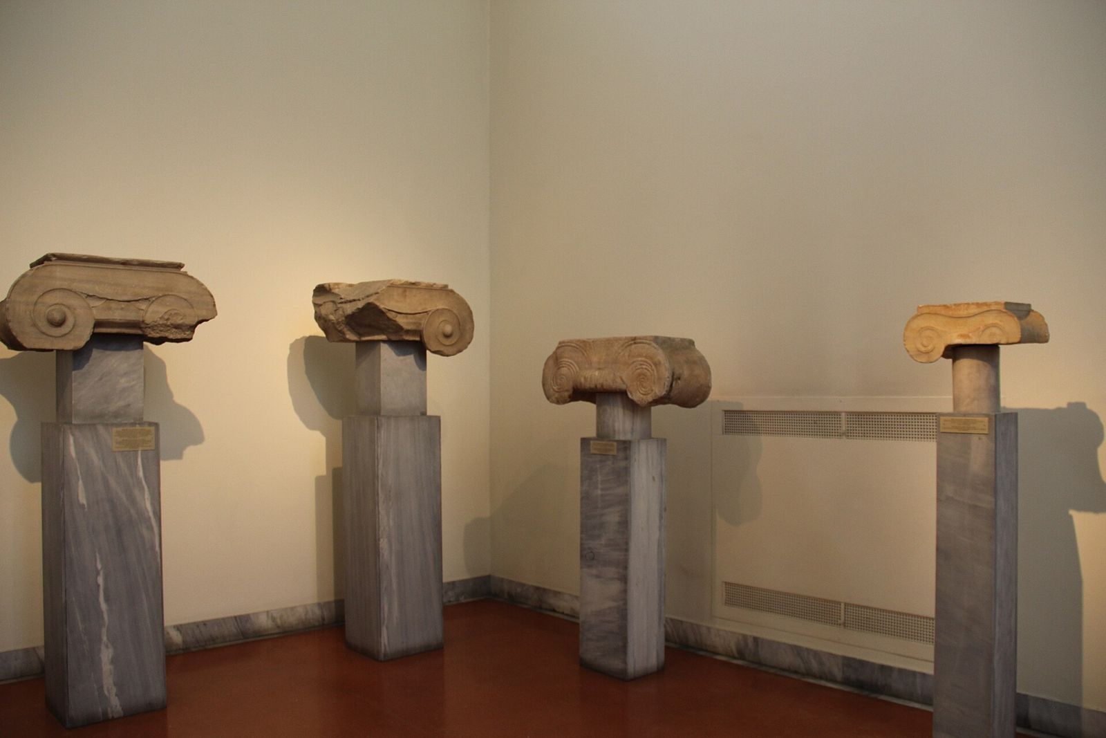 Four ancient marble column capitals on plinths in a gallery